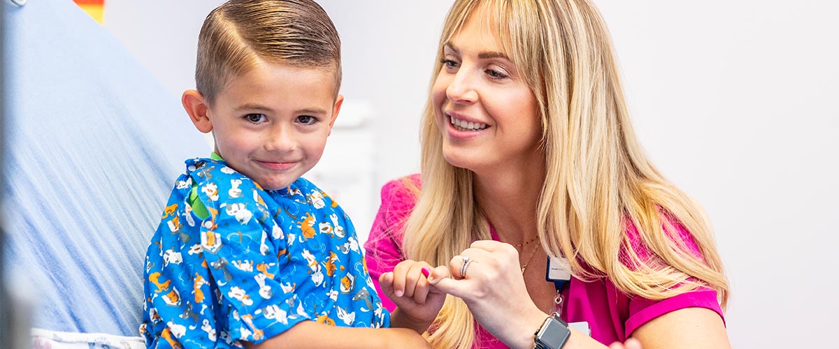 Little boy in blue gown on exam table and nurse holding his hand