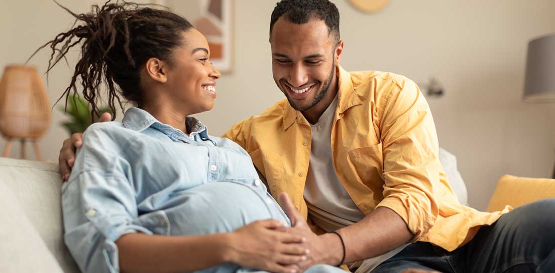 Happy african american couple expecting baby, pregnant black spouses embracing on couch at home. Smiling husband and wife resting in living room