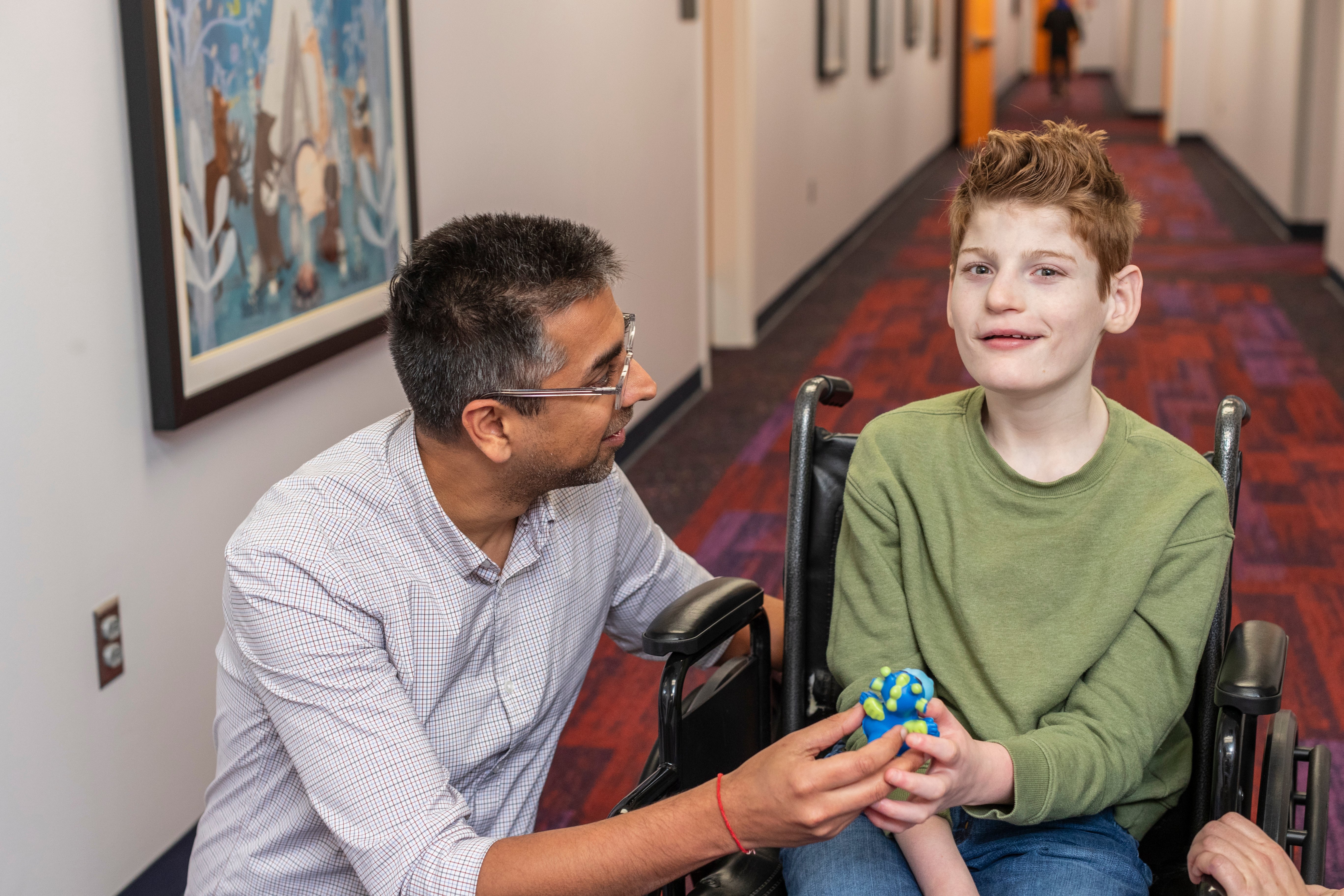 Young boy wearing a green shirt sitting in a wheelchair with doctor in glasses wearing a blue checkered shirt kneeling next to him