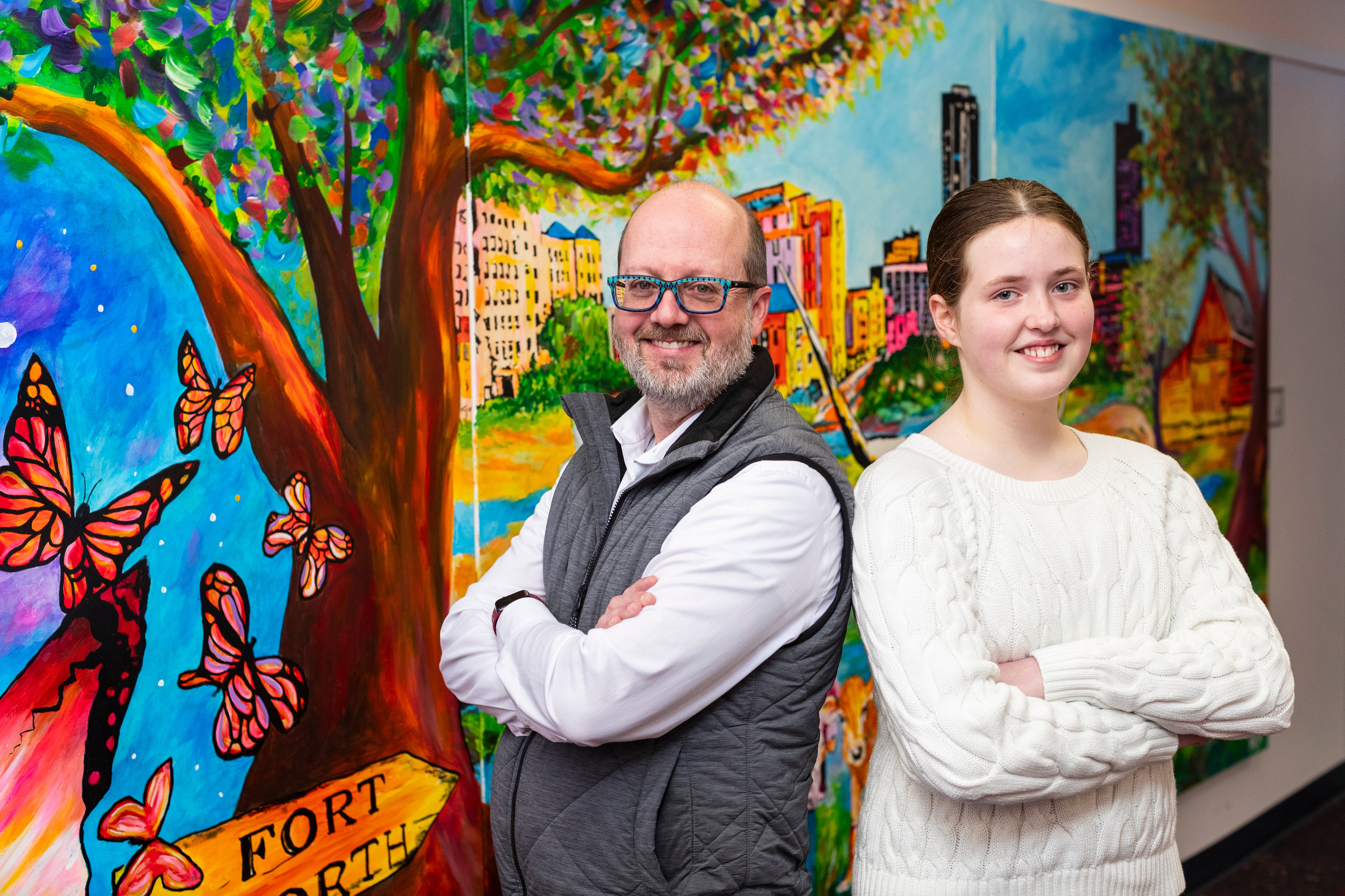 Doctor wearing blue glasses and a grey vest stands next to a young girl in a white sweater, they are in front of a colorful mural with butterflies